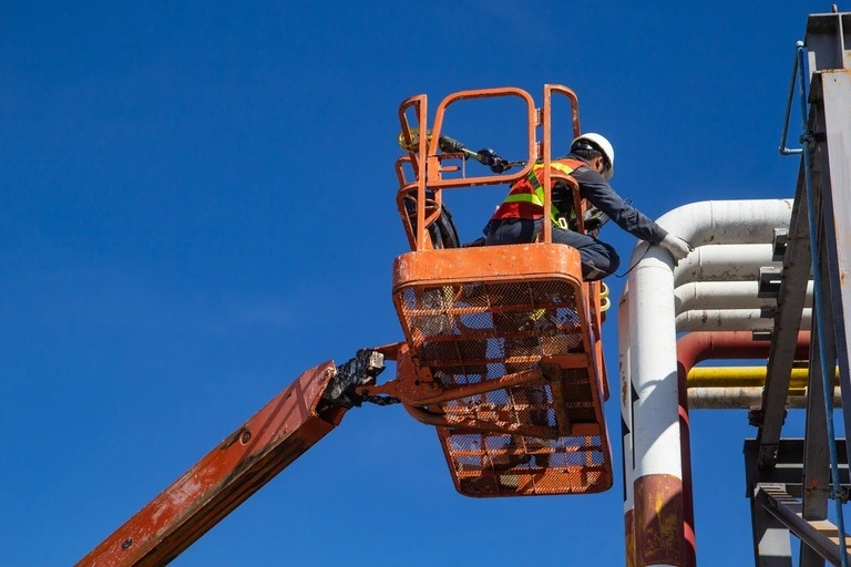 Male employee working up high in a boom lift inspecting the thickness of pipeline oil