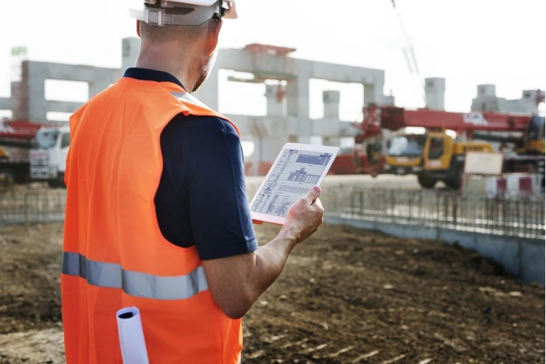 Construction contractor in an orange hi-vis jacket looking at site plans on his tablet with building site in the background
